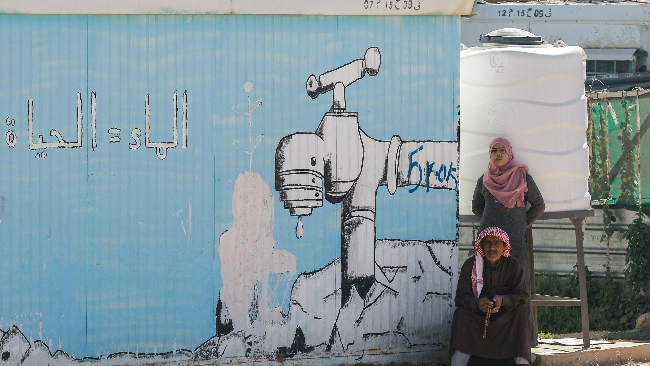 A woman stands behind a sitting man next to a cistern and a mural on a temporary shelter showing a dripping tap with a caption reading in Arabic "Water = Life", at the Zaatari refugee camp in northern Jordan.