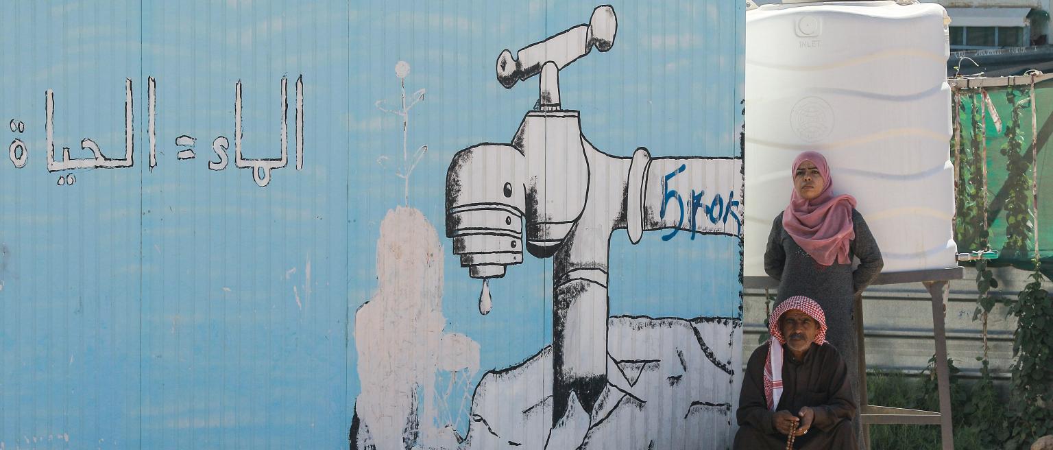 A woman stands behind a sitting man next to a cistern and a mural on a temporary shelter showing a dripping tap with a caption reading in Arabic "Water = Life", at the Zaatari refugee camp in northern Jordan.