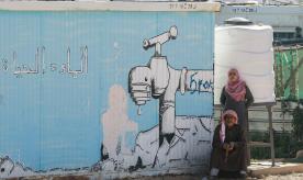 A woman stands behind a sitting man next to a cistern and a mural on a temporary shelter showing a dripping tap with a caption reading in Arabic "Water = Life", at the Zaatari refugee camp in northern Jordan.