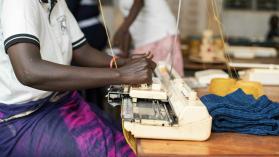 A person in a white shirt sitting at a brown wooden table, using a flat knitting machine