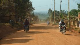 How distance affects public service delivery illustrated by locals driving motorbikes on a dusty road in Bunjako, Mpigi district, Uganda.
