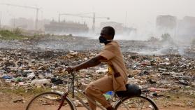 A person on a bicycle with a mask on cycling past a rubbish site, showing the need for urban waste management in African cities. 